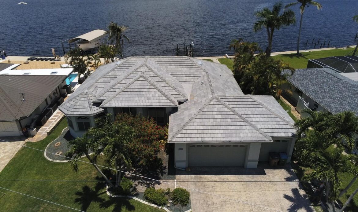 Aerial view of a waterfront residential home with a light colored tile roof, palm trees, and dock overlooking a lake in a coastal neighborhood
