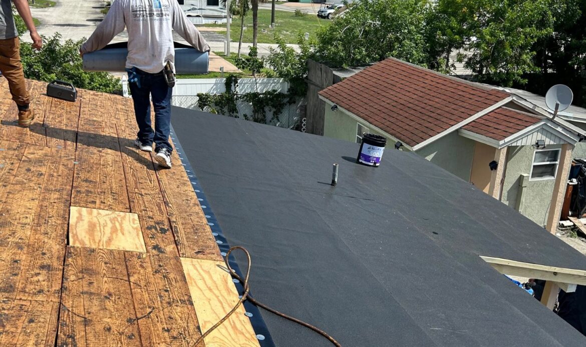 Two workers installing roofing material on a house, with one worker carrying a roll of roofing material on a bright sunny day